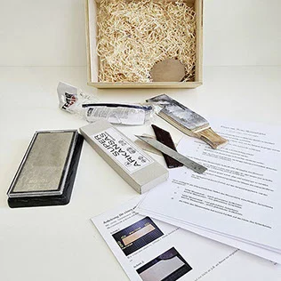 Inhalt der Michl-Box „Das Messer schärfen": Schleifstein, Messer, Abziehleder und Unterlagen vor der Holzkiste